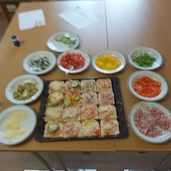 Ein Holztisch hat mehrere Teller mit verschiedenen Zutaten darauf. Auf dem Tisch befindet sich ein Tablett mit Pizzastücken und Schüsseln mit gehacktem Gemüse, Käse und Schinken.