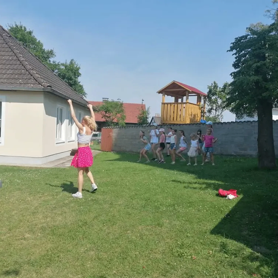 Eine Gruppe von Kindern spielt an einem sonnigen Tag draußen. Eine Frau führt sie bei einem Spiel an, während sie sich hinter einer Mauer aufstellen.