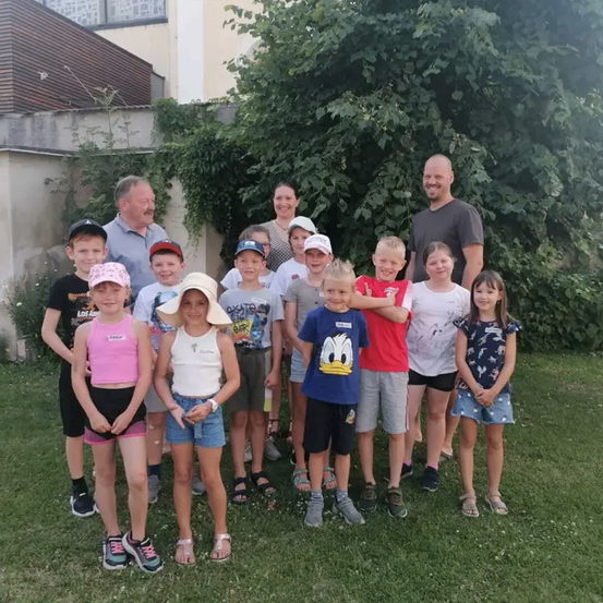 Eine Gruppe von Kindern und Erwachsenen steht in einem Grasbereich und lächelt für die Kamera. Sie tragen Hüte, T-Shirts und Turnschuhe. Sie stehen vor einem Gebäude mit Pflanzen und Bäumen.