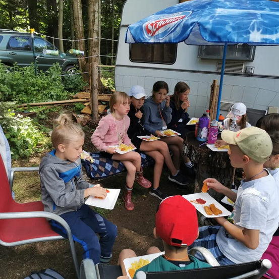 Eine Gruppe von Kindern isst zusammen im Freien unter einem blauen Regenschirm. Einige sitzen auf Stühlern, andere auf dem Boden.