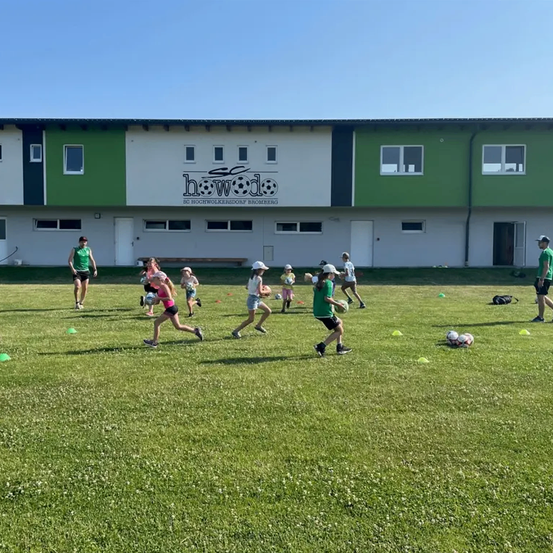 Kinder rennen auf einem Fußballplatz vor einem Gebäude mit dem Logo von SC Hochwolkersdorf Bromberg.