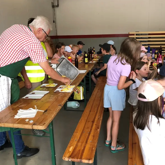 Eine Gruppe von Menschen in einem Saal, ein älterer Mann serviert Essen auf einem Tisch, während andere zusehen.