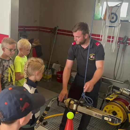 Ein Feuerwehrmann demonstriert die Verwendung einer Schlauchtrommel für eine Gruppe von Kindern in einem Raum mit weißen und roten Wänden.