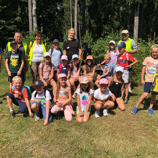 Eine Gruppe von Kindern und Erwachsenen posiert für ein Foto in einem Grasbereich, einige stehen hinter einer Bank, und es gibt Bäume im Hintergrund.