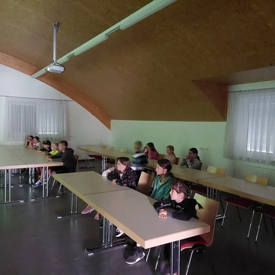 Kinder sitzen um Tische in einem Klassenzimmer mit hölzernen Bögen an der Decke, grünen Wänden und weißen Vorhängen an den Fenstern.