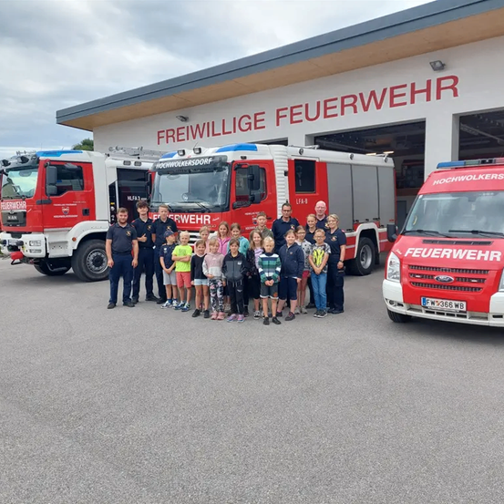 Eine Gruppe von Feuerwehrleuten und Kindern posiert vor der Feuerwache mit zwei roten Feuerwehrwagen.