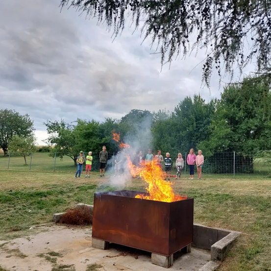 Eine Gruppe von Kindern beobachtet ein Feuer in einem Metallbehälter, das hell in einem Außenfeld brennt.