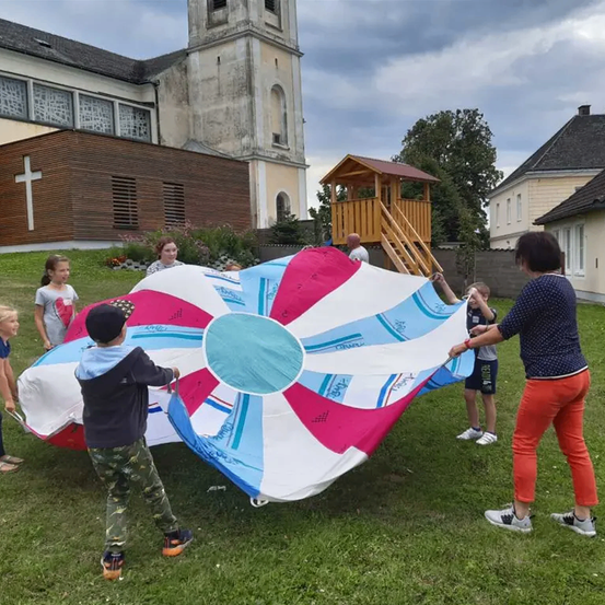 Kinder und Erwachsene spielen mit einem bunten Fallschirm auf dem Rasen vor einer Kirche.