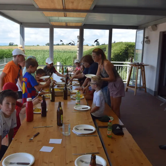 Eine Gruppe von Kindern und Erwachsenen sitzt um einen langen Tisch mit Tellern und Flaschen und isst. Der Tisch steht auf einem Balkon mit einer Glasfront. Der Balkon hat Blick auf ein Grasfeld.