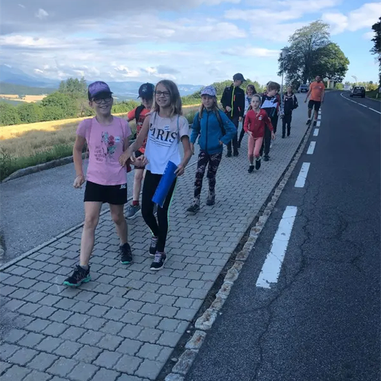 Eine Gruppe von Kindern und Erwachsenen, die auf dem Gehweg einer Straße gehen, mit einem Grasfeld und Bäumen im Hintergrund.
