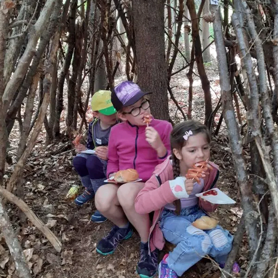 Drei Kinder sitzen in einer Hütte aus Ästen im Wald. Zwei von ihnen essen Hot Dogs, während das andere sie ansieht.