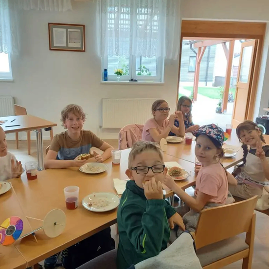 Eine Gruppe von Kindern sitzt um einen Tisch in einem Raum. Sie essen und trinken. Der Tisch ist mit Tellern, Bechern und einem bunten Rad bedeckt. Der Raum hat eine weiße Wand mit einem Bilderrahmen und einem Fenster mit Blick nach draußen.
