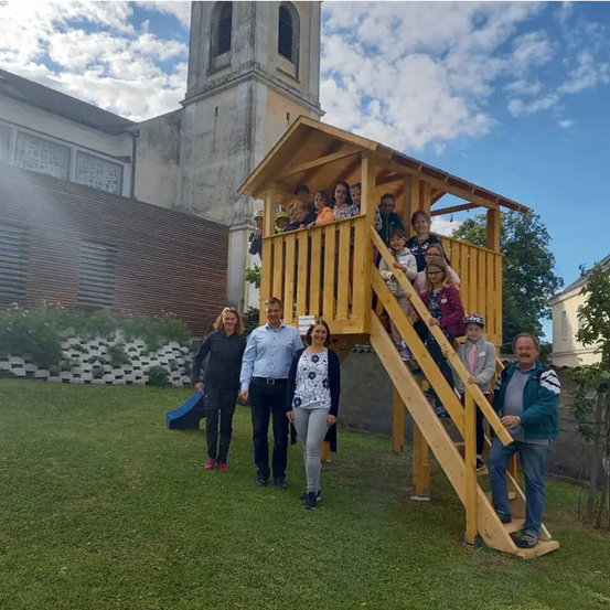 Gruppenfoto von Erwachsenen und Kindern in einem Spielplatz mit einem Holzhaus, einer blauen Rutsche und einem grünen Rasen. Dahinter steht ein Gebäude mit Turm und Mauer.