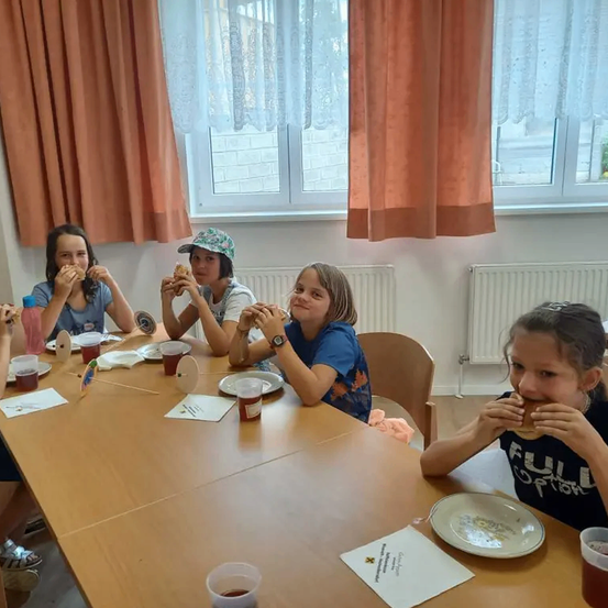 Vier Mädchen sitzen an einem Tisch in einem Raum und essen Sandwiches. Ein Mädchen auf der rechten Seite trägt ein T-Shirt mit dem Text FULL OPTION. Auf dem Tisch stehen Teller, Tassen und Servietten. Hinter ihnen befinden sich Heizkörper und Vorhänge.