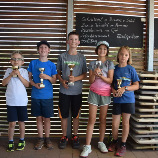 Fünf Jugendliche stehen gemeinsam vor einer Holzwand mit einer Tafel und halten ihre Trophäen.