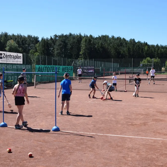 Eine Gruppe junger Menschen spielt Tennis auf einem Tennisplatz. Im Hintergrund sind Bäume zu sehen.