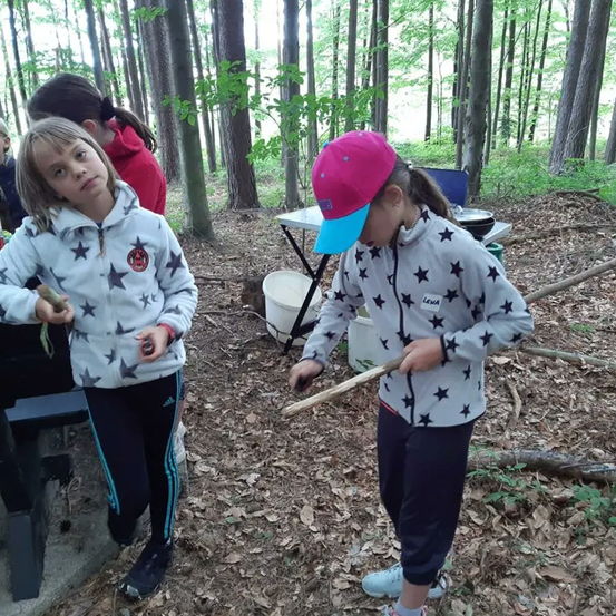 Zwei Mädchen, eines mit einer rosa Mütze, stehen in einem Wald und halten Stöcke in der Hand und schauen sich etwas auf dem Boden an.