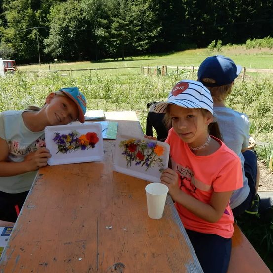 Drei Kinder sitzen draußen an einem Tisch. Sie haben Blumen auf ihrem Tisch und lächeln in die Kamera.