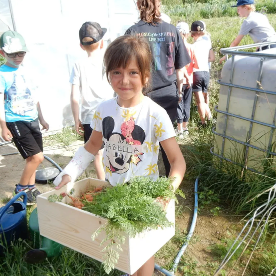 Ein Mädchen mit einem Gips auf dem linken Arm hält eine Box mit frisch geernteten Karotten und Petersilie. Sie lächelt und steht zwischen anderen Kindern in einem Gartenbereich. Hinter ihr stehen einige andere Kinder, und einige Erwachsene stehen an der Seite. Ein Metallwassertank ist an der Seite aufgestellt.