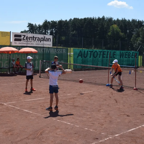Vier Kinder spielen Tennis auf einem Platz, wobei ein Mann Schläger und Ball hält. Im Hintergrund stehen zwei Regenschirme.