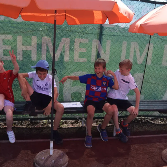 Vier Jungen sitzen auf einer Bank unter einem orangefarbenen Regenschirm. Sie lächeln und scheinen Spaß zu haben. Ein Junge hat einen Baseballschläger neben sich.