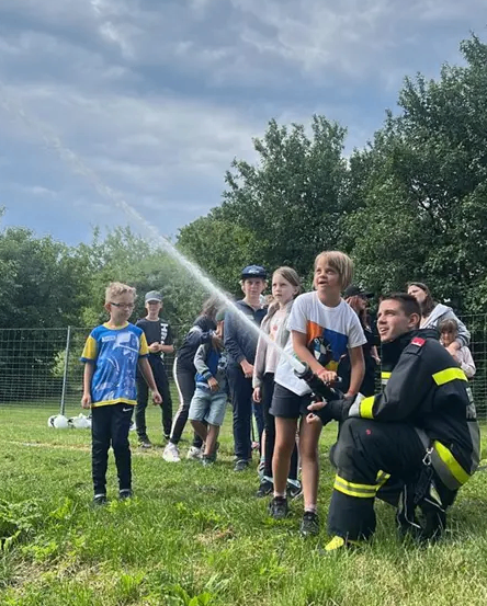 Feuerwehrmann zeigt Kindern, wie man einen Wasserschlauch benutzt, in einem Feld mit Bäumen und einem Zaun im Hintergrund.