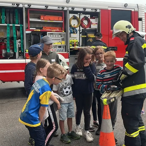 Ein Feuerwehrmann spricht mit einer Gruppe von Kindern, die vor einem Feuerwehrwagen stehen, dessen Tür offen ist und verschiedene Ausrüstungsgegenstände im Inneren zeigt.