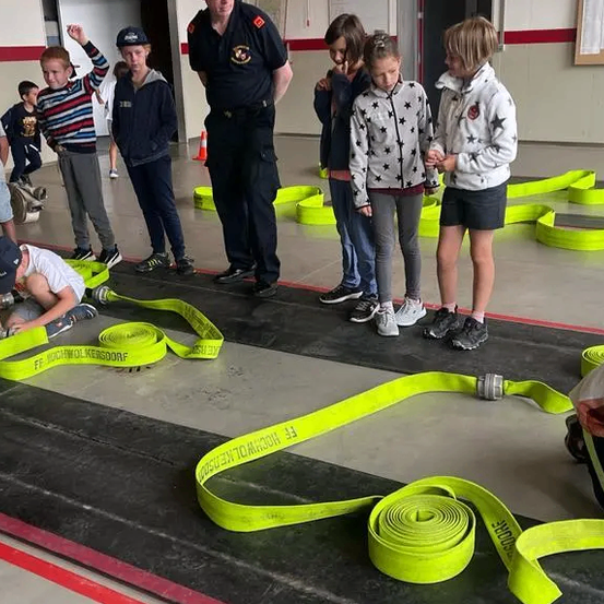 Eine Gruppe von Kindern und Erwachsenen befindet sich in einem Gebäude mit einem Feuerwehrmann, sie stehen auf einem schwarz-gelb gestreiften Boden mit gelben Bändern und scheinen einige Aktivitäten durchzuführen.