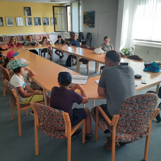 Eine Gruppe von Kindern und Erwachsenen sitzt um einen langen Tisch in einem Klassenzimmer. Das Klassenzimmer hat gelbe Wände, eine weiße Tür und mehrere gerahmte Bilder. Die Kinder tragen Hüte und Turnschuhe.