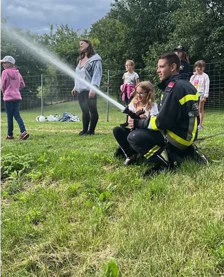 Ein Feuerwehrmann demonstriert einer Gruppe von Kindern die Verwendung einer Feuerwehrspritze auf einer Wiese.