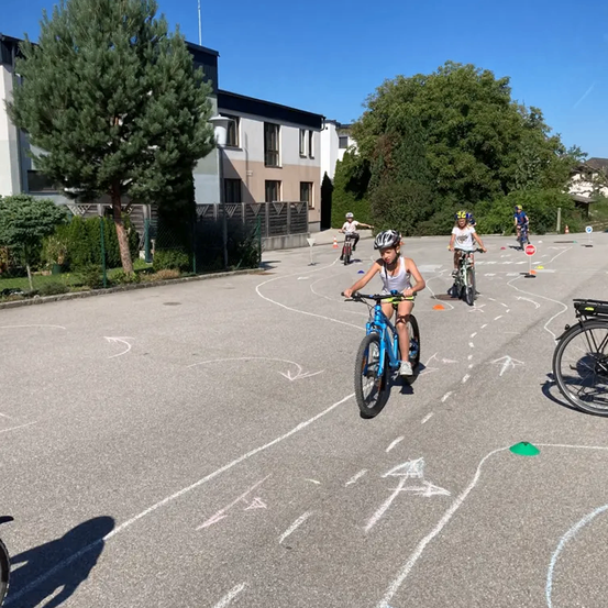 Vier Kinder fahren auf einer Straße mit weißen Markierungen und grünen Hütchen mit Fahrrädern. Das Kind vorne trägt einen Helm und Turnschuhe. Es gibt Bäume, Büsche und Gebäude am Straßenrand.