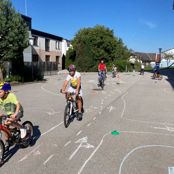 Kinder fahren auf einem Parkplatz mit einem Mann, der sie unterrichtet, auf Fahrrädern. Der Parkplatz ist mit Pfeilen und Kreisen bemalt. Im Hintergrund befinden sich Bäume und Gebäude.