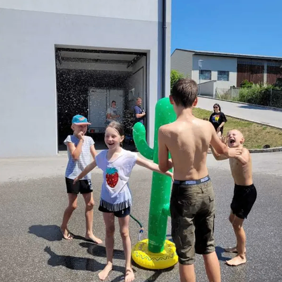 Vier Kinder spielen auf der Straße mit einem Wasserbrunnen. Sie lächeln und scheinen das kühle Wasser zu genießen.