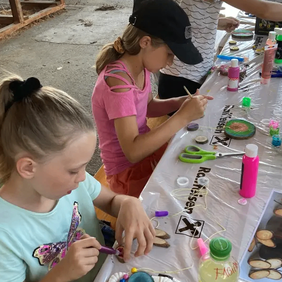 Drei Mädchen malen an einem Tisch mit Bastelutensilien. Ein Mädchen trägt ein rosa Shirt und das andere ein blaues. Sie verwenden Pinsel und andere Werkzeuge, um ihre Kunstwerke zu gestalten.