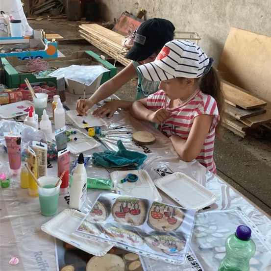 Zwei Mädchen sitzen an einem Tisch und malen auf Holzscheiben. Verschiedene Malutensilien und Farben sind auf dem Tisch. Hinter ihnen sind Holzbretter und Kisten.