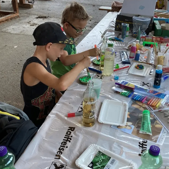 Zwei Kinder malen auf einem Tisch mit verschiedenen Malutensilien wie Flaschen, Pinseln und Papieren.