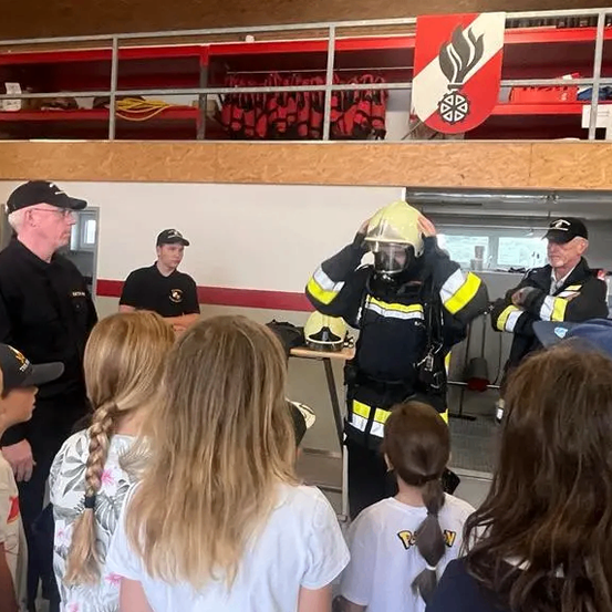Ein Feuerwehrmann demonstriert Kindern einen Schutzhelm. Die Kinder schauen den Feuerwehrmann an.
