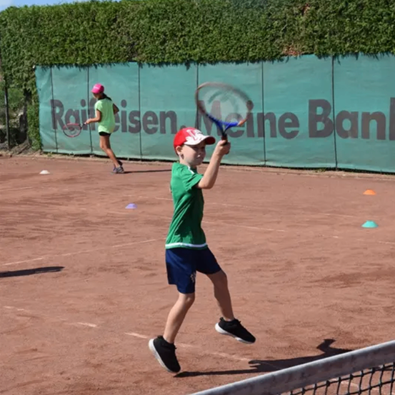 Ein Junge schlägt einen Tennisball mit einem Tennisschläger auf einem Tennisplatz, eine Mädchen ist im Hintergrund zu sehen.