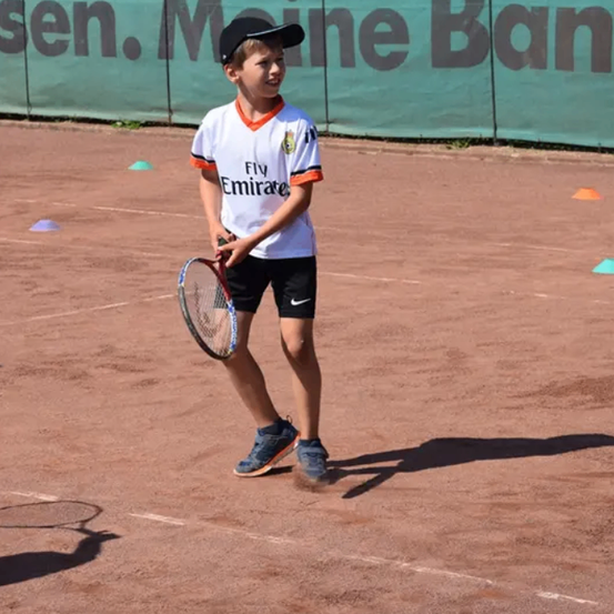 Ein Junge in einem weißen T-Shirt mit orangefarbenen Akzenten spielt Tennis auf einem Sandplatz.