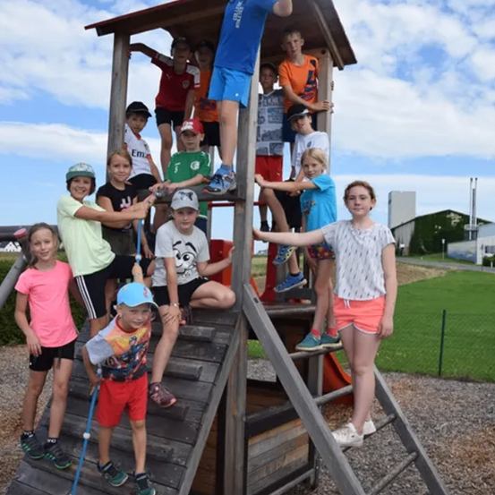 Eine Gruppe von Kindern versammelt sich um ein hölzerne Spielhaus. Einige stehen auf der Treppe, andere im Inneren des Spielhauses, und ein Junge steht obenauf.
