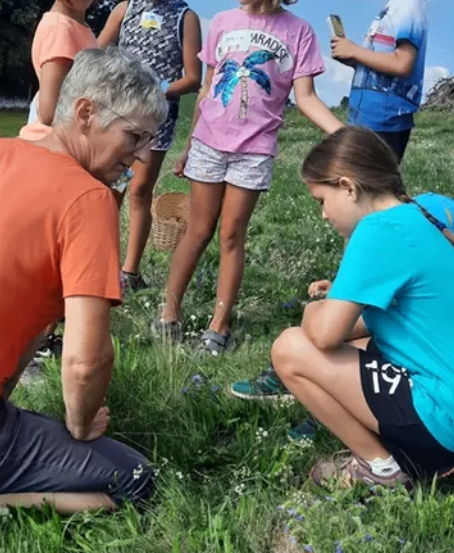 Eine Gruppe von Kindern und einem erwachsenen Mann befindet sich auf einem Rasen, einige knien, einige stehen und einige halten einen Korb. Der erwachsene Mann hockt und schaut auf etwas am Boden.