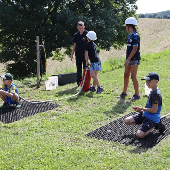 Vier Kinder sind an einem sonnigen Tag draußen. Zwei Kinder knien und halten einen Schlauch, während ein weiteres Kind steht und zusieht.