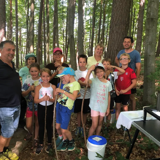 Gruppe von Erwachsenen und Kindern, die mit Stöcken in einem Wald stehen und in die Kamera lächeln. Ein Tisch mit einer weißen Tischdecke ist im Hintergrund.
