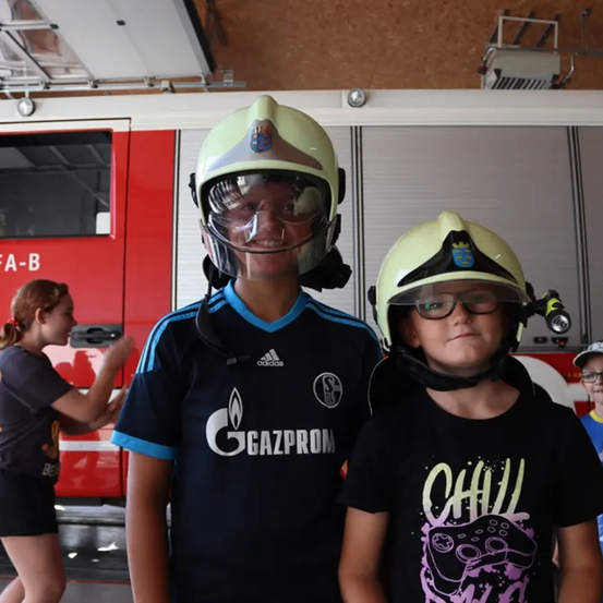 Zwei Jungen mit Helmen in einer Feuerwache mit einem Feuerwehrwagen dahinter.