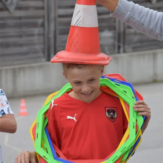 Ein Junge mit einem roten Shirt, einem Puma-Logo, einem bunten Hula-Hoop-Reifen und einem orangefarbenen Verkehrskegel auf dem Kopf lächelt.