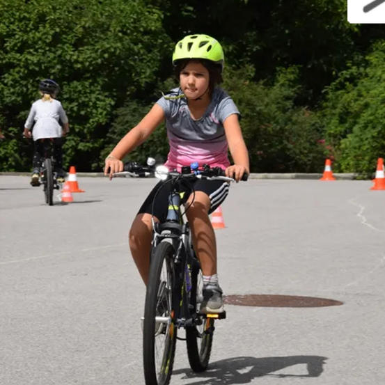 Ein Mädchen mit einem grünen Helm fährt auf einer asphaltierten Fläche mit einem Fahrrad. Orangefarbene Verkehrskegel sind um sie herum aufgestellt. Ein weiteres Mädchen fährt hinter ihr ein Fahrrad, und Bäume sind im Hintergrund.