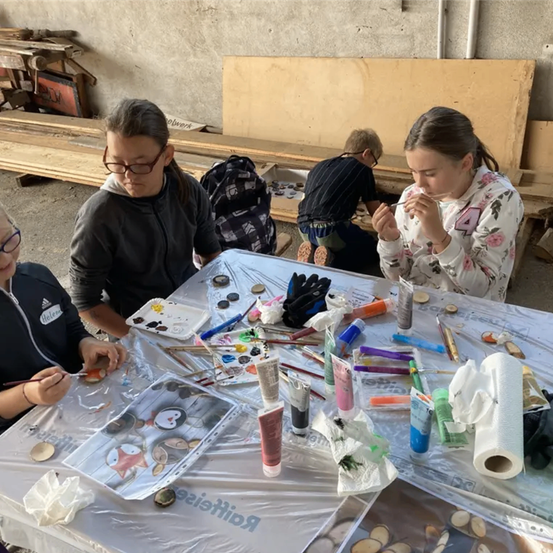 Drei Mädchen malen auf einem Tisch in einem Außenbereich. Ein Mädchen hält einen Pinsel und malt auf ein Papier.