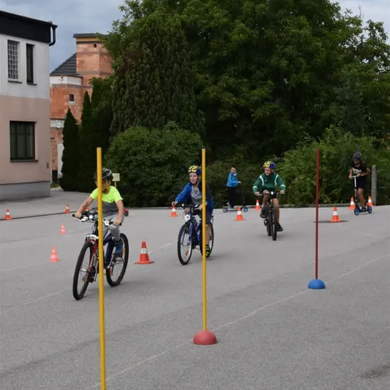 Fünf Kinder fahren auf einem Parkplatz mit Fahrrädern, umgeben von orangefarbenen und gelben Hütchen. Sie tragen Helme und Turnschuhe.
