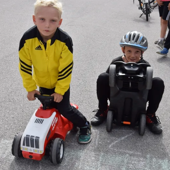 Zwei junge Jungen sind draußen auf einer asphaltierten Straße. Ein Junge fährt ein Spielzeugauto, während der andere Junge auf einem Spielzeuglastwagen sitzt. Beide Jungen tragen Jacken und Turnschuhe.