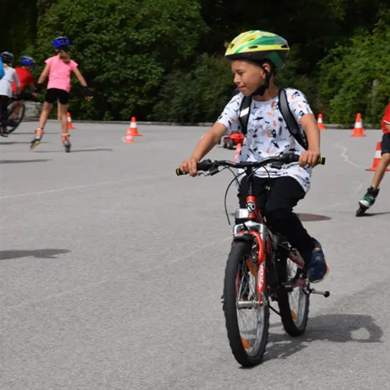 Ein junger Junge fährt ein Fahrrad auf einem asphaltierten Bereich mit mehreren anderen Kindern. Sie sind von Verkehrskegeln und grünen Bäumen umgeben.